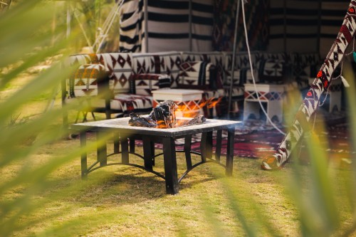 A scene of an outdoor seating area featuring a traditional tent with furniture adorned with Arabic motifs and patterned fabrics. In the foreground, there is a lit fire pit on a metal table, surrounded by a grassy ground and a backdrop with colorful tribal woven patterns.