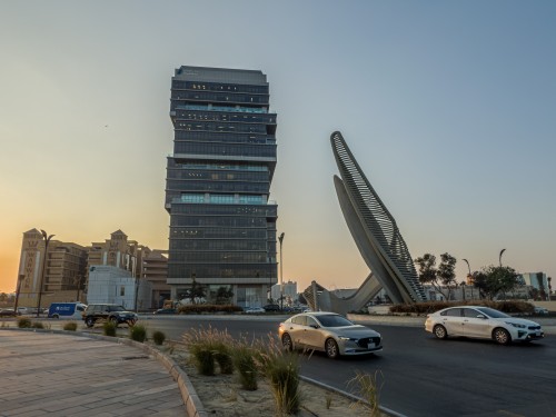 صورة لشارع في مدينة على وقت الغروب, يظهر فيها مبنى زجاجي حديث ذو تصميم معماري فريد يتكون من عدة طوابق بشكل متدرج. بجوار المبنى توجد نصب تذكاري كبير ذو شكل منحني. السيارات تسير في الطريق, وتظهر بعض النباتات في مقدمة الصورة.

-