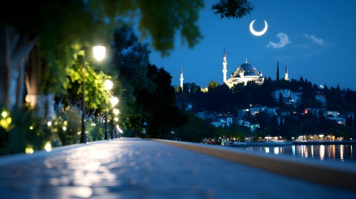 A nighttime view of a large mosque illuminated with white lights, featuring tall minarets, set in the background of a city overlooking a calm lake. The sky is a deep blue with a bright crescent moon above, while streetlights warmly illuminate the pathway by the lakeside, with trees and greenery scattered along the sides.