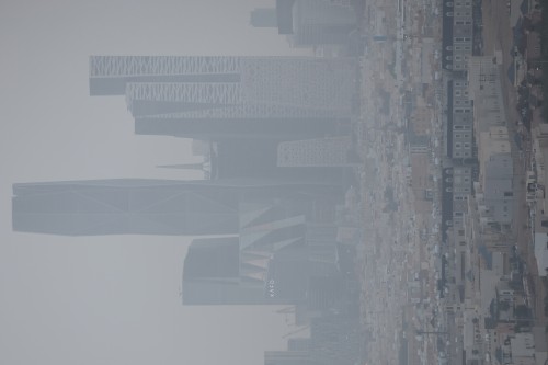 A general view of Riyadh\'s skyline featuring several modern skyscrapers with distinctive architectural designs. The image shows multiple buildings aligned in a hazy scene. The setting appears urban and developed with multi-story buildings, roads, and streets.