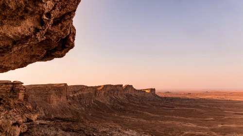 A natural landscape featuring a stretch of rocky cliffs in a desert area under a light blue sky during sunrise or sunset. The prominent mountain rocks extend dramatically across the scene, showing gradients of brown and yellow blending with the soft sunlight.