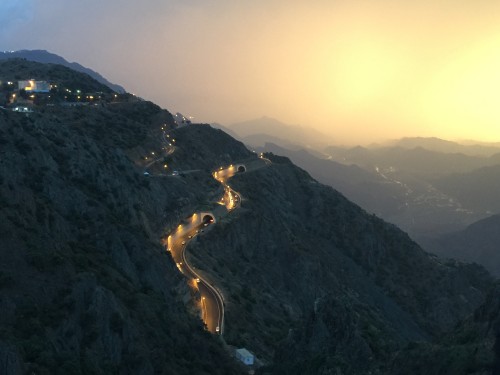 A view of a winding mountain road lit by street lamps in the evening, nestled between mountainous terrain. The sky in the background displays shades of orange and gold, with silhouette-like mountain ranges extending into the distance. Some houses and lights are visible in the upper left corner of the image.