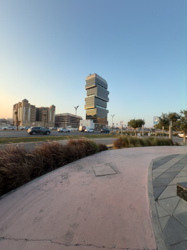 مبنى حديث بتصميم فريد يظهر مكوناً من عدة طوابق متكدسة فوق بعضها, يقع بجواره مباني تقليدية أخرى تحت سماء زرقاء صافية. يمكن رؤية السيارات على الطريق وأعشاب تزيينية على الرصيف القريب. الأفق يوحي بجو هادئ في وقت الغروب.

-