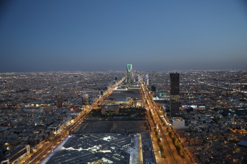 Aerial view of Riyadh\'s skyline at night, showcasing a long illuminated road running through the city leading to the Kingdom Tower, distinguished by its unique design and green lighting. Surrounding are buildings and lit streets, reflecting the urban landscape of the city.