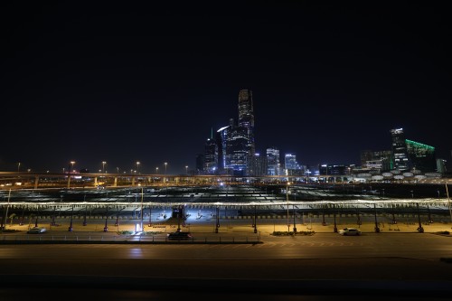 A nighttime image of a modern cityscape with a skyline illuminated by the lights of tall buildings. Glass towers are lit up in blue and green. In the foreground, a wide road and a parking lot under streetlights. On the right, a building with bright green lights resembles a contemporary structure.