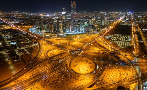 Aerial night view of a city illuminated with bright lights. A complex network of highways and intersections is prominent in the foreground, surrounded by tall buildings and skyscrapers in the background, sparkling with lights. The lighting details highlight the entire scene.