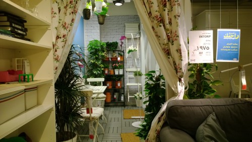 The image shows an interior section of a furniture store featuring a seating area. There is a grey sofa with cushions, and floral patterned curtains hanging on each side. The back of the image displays a white table and chairs surrounded by various potted plants. The walls are decorated with white brick, and the shelves hold books and small decorations. There are also signs hanging with product information.