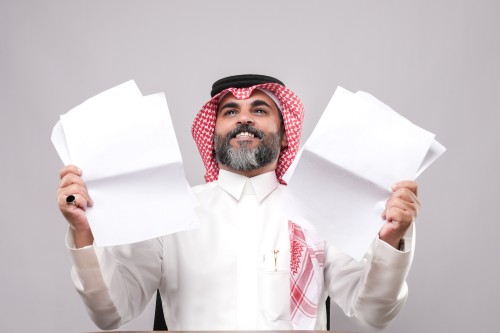 رجل يرتدي الزي التقليدي الخليجي ويمسك بأوراق بيضاء بيده, يبدو سعيدًا.