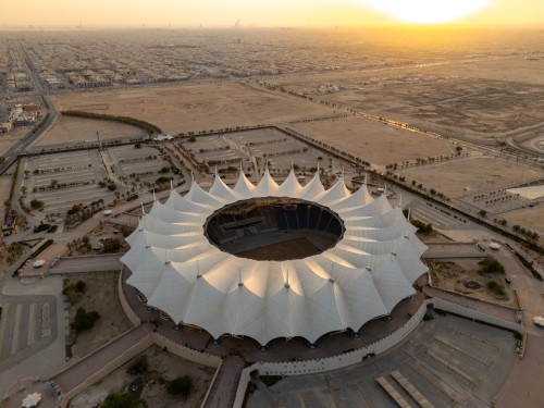 An aerial view of King Fahd Stadium in Riyadh, Saudi Arabia. The stadium features a distinctive design resembling a large white tent surrounded by vast expanses of land and empty parking lots. The city extends into the horizon with the sunrise visible in the upper right corner of the image, adding a warm glow to the scene.
