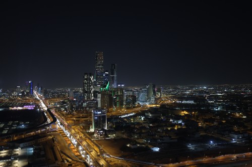 Night view of Riyadh city in Saudi Arabia, showcasing the city skyline adorned with lights. Tall illuminated buildings and the famous tower with its triangular design are visible. The highways on the ground are lit, with cars moving along them.