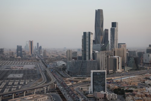 Aerial view of Riyadh, the capital of Saudi Arabia, showcasing modern buildings and skyscrapers with diverse architectural designs. A tall tower dominates the image alongside other multi-story buildings. Busy highways filled with vehicles run through the scene, and the King Abdullah Financial District is notably featured.