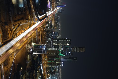 A nighttime image of Riyadh city in Saudi Arabia, showing a brightly lit highway with streaks of lights from moving cars in heavy traffic. Skyscrapers illuminate the skyline, with the Kingdom Tower prominently visible in the background, lit with multicolored lights.