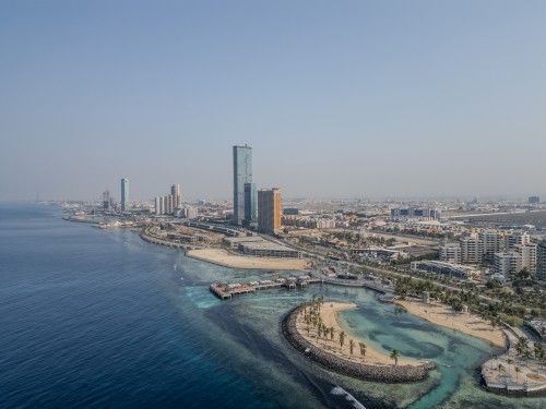 An aerial view of a modern waterfront in a city located on the Red Sea coast, featuring a cluster of tall buildings and hotels. Palm trees are lined along the sandy beach, with a stretch of roads and tourist facilities. The clear blue waters extend to the horizon under a clear sky.