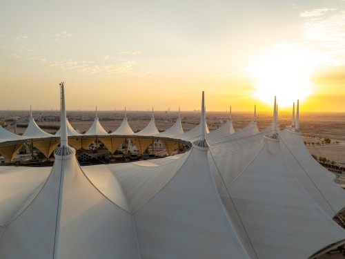 Aerial view of large white canopies at King Fahd Stadium in Riyadh, Saudi Arabia, designed in the shape of traditional tents. The sunset bathes the scene in golden hues, with the city and a few soft clouds visible in the background.