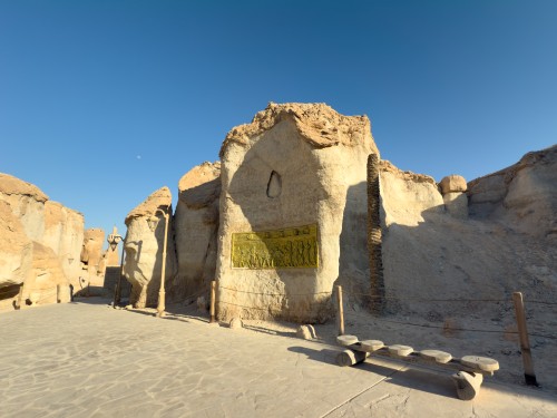 منظر لجدران صخرية طبيعية في موقع أثري تحت سماء زرقاء صافية. يوجد نقش نحاسي على الجدار الأوسط, ويظهر ظل عمود إنارة قديم على الأرض المعبدة. علامات الطريق مصنوعة من الحبال وجذوع الأشجار, مما يضيف لمسة تقليدية للمكان.

-