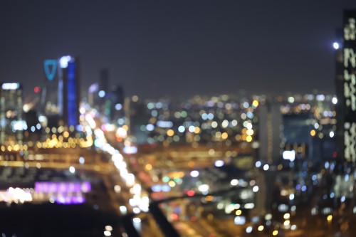 A nighttime cityscape with illuminated buildings and streets, appearing blurred. Tall buildings with lights are visible in the background, creating a bokeh effect that reflects the city lights.