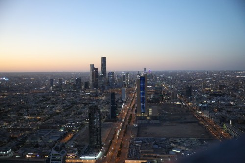 Aerial view of Riyadh in Saudi Arabia at sunset, featuring modern skyscrapers with sparkling lights. The sky gradually fills with an orange hue as light envelops the city. Buildings are scattered widely in the background.