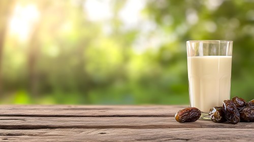 A glass of milk placed on a wooden table with a pile of dates beside it. The background features a blurred natural view, adding a sense of calmness and nature.