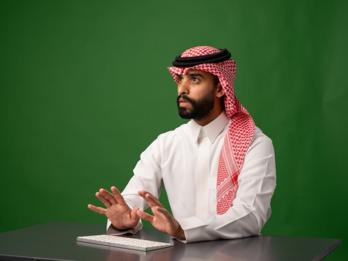 رجل عربي يرتدي الزي الخليجي التقليدي يجلس أمام لوحة مفاتيح مع خلفية خضراء.