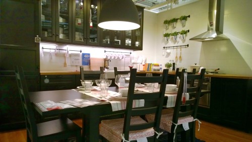 An image of a modern kitchen featuring a dark wooden dining table surrounded by several chairs. The table is set with dishware, including plates and glass cups. There is an overhead cabinet with glass doors displaying shelves of dishes. A wall-mounted arrangement includes small potted plants and hanging kitchen utensils. A metallic range hood is above the cooking area in the corner.