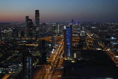 A view of Riyadh city in the evening, showcasing the skyline filled with towers and tall buildings, with a central tower illuminated in blue. The city lights glitter throughout, reflecting the vibrant nightlife of the city.