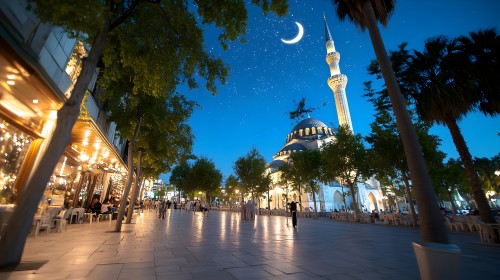 Image of a city under moonlight, featuring an Ottoman-style mosque with a large dome and a tall, illuminated minaret. In front of the mosque are paved streets with trees, outdoor seating, and beautifully lit cafes. The sky is starry with a bright crescent moon.