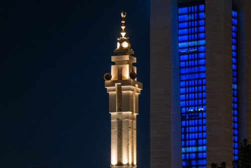 Nighttime image of a section of a mosque minaret illuminated with warm white lights, featuring a small dome and crescent at the top. Loudspeakers are visible on the sides. In the background, a building with bright blue lights in its windows can be seen, creating a contrast between the blue and white lighting.