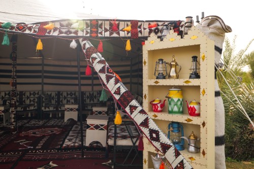 The image shows a traditional tent adorned with bright colors and geometric patterns. A wooden shelf holds traditional metal pots, including teapots and coffee pots, alongside lanterns. The tent interior is lined with traditional textiles featuring heritage designs, accompanied by traditional Arabian seating arrangements.