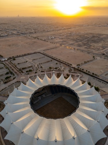 Aerial view of King Fahd Stadium in Riyadh, Saudi Arabia, at sunset. The stadium is characterized by its unique circular design and distinctive white roofs. In the background, the cityscape of Riyadh extends into the desert with the sunset casting a warm golden hue across the sky.