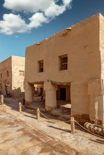 A traditional two-story mud building with small exterior columns. The building features wooden windows and large doors, designed in a traditional Arabic architectural style. The blue sky with a few white clouds is visible in the background, giving the image a sense of calm and tranquility.