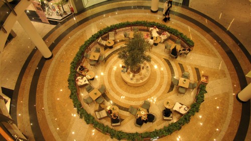 Aerial view of a circular seating area in a shopping mall, featuring tables and chairs arranged around a planted tree in the center. People are seated around the tables, some wearing traditional attire. The area is surrounded by green plants with a marbled floor design.