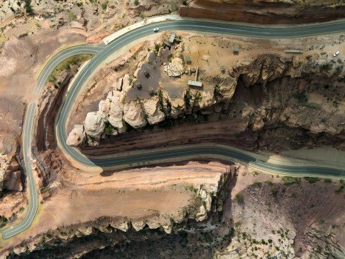 An aerial view of a winding road passing through a rocky mountainous area. The rocks display varying shades of brown and red, surrounded by cliffs and rocky walls. Sparse greenery is visible along the road, with one car traversing it.
