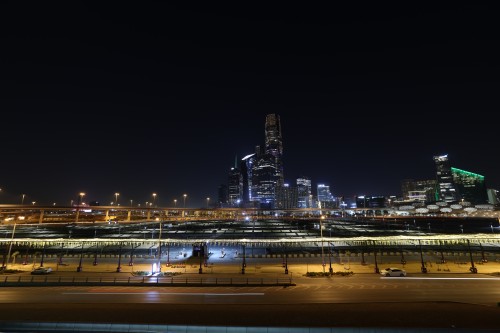 A nighttime view of a modern city with brightly lit skyscrapers on the horizon. In the foreground, there are empty parking lots and roads with street lighting. Several towers of varying heights are prominent, some featuring colorful lights.