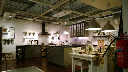 An image of a modern kitchen with stylish interior design. The kitchen features white tiled walls and grey cabinets, with large countertops holding kitchen tools and utensils. Green plants are placed on the shelves, and framed pictures hang on the wall. Pendant lights hang from the ceiling, and chairs are placed around a side table. The kitchen floor is dark wood, and on the right, there is a red fire extinguisher.