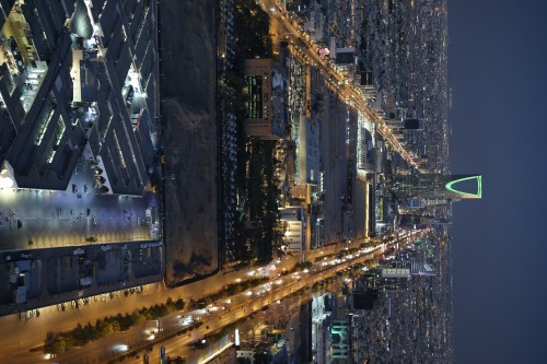 A night aerial view of a bustling city in Saudi Arabia, showcasing multiple illuminated roads and tall buildings, including the prominent Kingdom Tower with its distinctive green lighting, highlighting the modern city skyline.