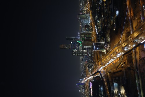 A nighttime image of Riyadh, Saudi Arabia, showing the city skyline illuminated with lights from tall buildings and busy streets filled with cars. The Kingdom Tower is clearly visible with lights surrounding it, along with various roads and bridges lit up at night. The dark sky accentuates the bright city lights.