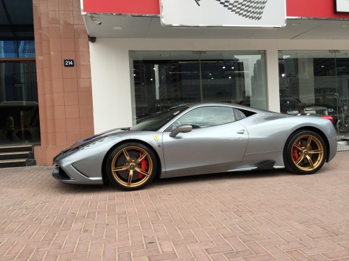 Image of a luxury silver sports car with gold wheels on a brick pavement. The car is parked in front of a modern building with a glass facade, with city landmarks visible in the background.