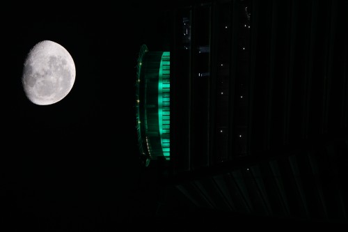 An image showing a bright full moon on the left, and on the right, part of a building illuminated with green lights. The visible section of the building has a modern design and contrasts with the dark night sky.