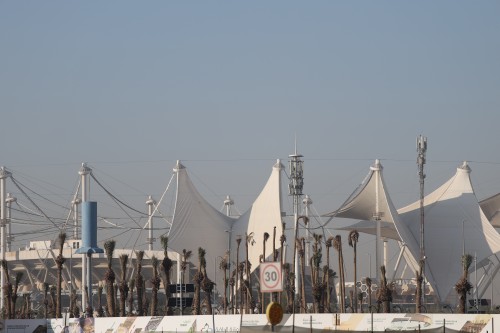The image shows large white structures resembling tents, which are part of a modern architectural design. In front, there are several palm trees and some metal towers, along with a sign indicating a speed limit of 30 km/h. The sky is clear, suggesting an urban setting.