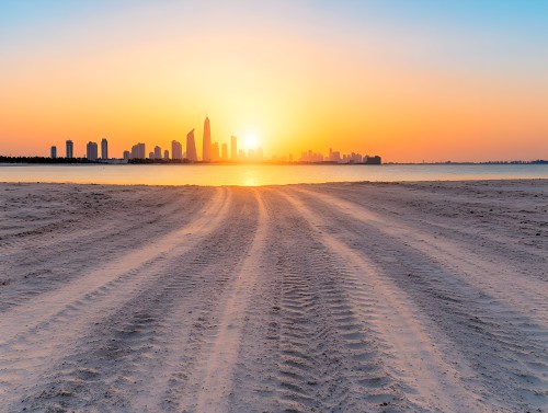 A sunset view over a modern city skyline with several skyscrapers. The foreground shows sandy tracks from vehicle tires leading to calm waters. The sky is filled with shades of orange and blue during the sunset.