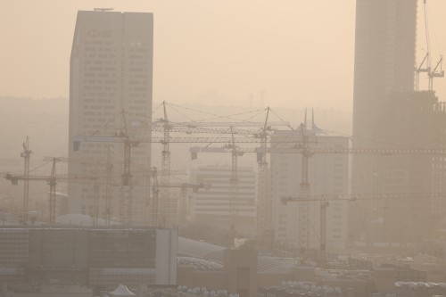 A cityscape under construction featuring several tower cranes in the foreground. Modern buildings are distributed in the background as a golden light mingles with dust in the air, giving a hazy appearance to the scene. A contemporary tower with a bank logo, alongside other high-rise buildings, indicates rapid urban development.