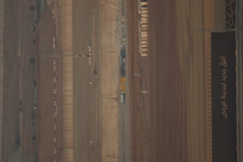 An aerial view of a construction site showing a long road stretching through the desert with parts of equipment and temporary structures. On the right side, there is a black barrier with Arabic writing that reads \"New horizons for the city of Riyadh.\" The ground is surrounded by light brown dirt and vast sandy areas.