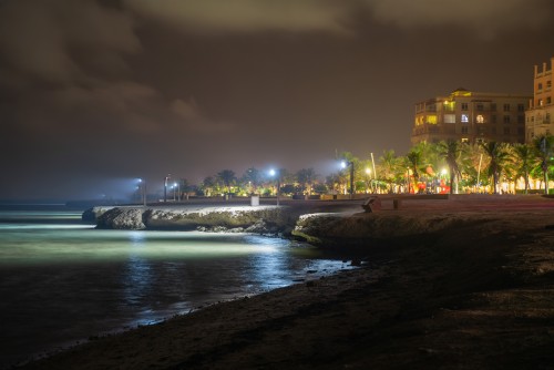 A night view of a seaside with street lights reflecting on the water. In the background, illuminated buildings, palm trees, and a large Ferris wheel are visible. The sky is dark with some clouds. The beach appears calm and deserted.