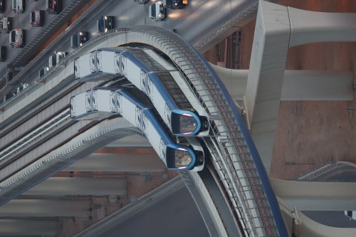 An aerial view of two trains moving in opposite directions on an elevated track next to a busy highway with cars. Each train is blue and white and travels on a curved track. The highway is crowded with cars moving in parallel lines beneath the trains.