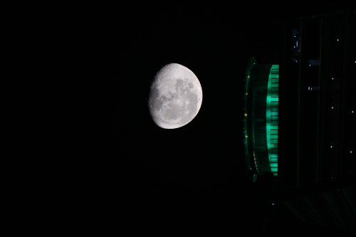 An image of the moon in the dark night sky, clearly visible with its white and gray details against the surrounding black sky. On the right side of the image, part of a building lit in green is visible.