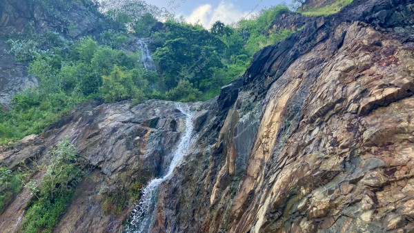 صورة لشلال صغير يتدفق عبر الصخور الكبيرة في جبل مغطى بالنباتات الخضراء والأشجار الكثيفة. يظهر في الصورة مزيج من الصخور البنية والرمادية مع المياه المتدفقة عبر الجبل في وسط الطبيعة الخصبة.