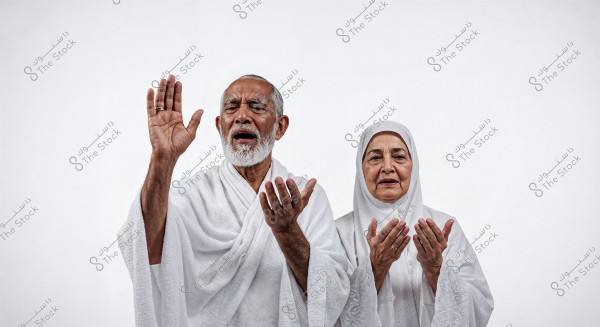 An image of an elderly man and woman wearing white Ihram clothing. The man raises his right hand in a gesture of prayer, while his left hand is open beside the woman, who wears a white headscarf. Both appear in a reverent posture against a white background.