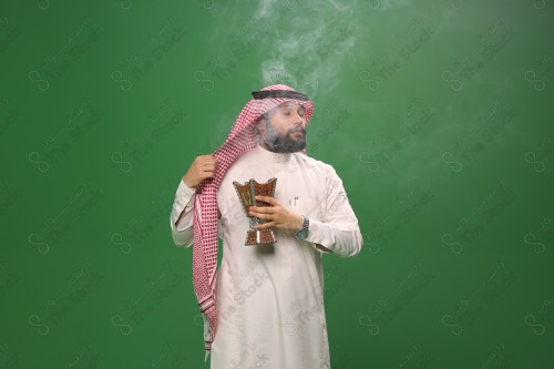 رجل سعودي يقف على خلفية خضراء يرتدي الثوب و الشماغ، يقوم بعمل حركات بيديه ، يبخر نفسه بالطيب و العود، احتفالات العيد