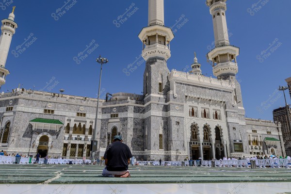 A man sits in contemplation in the courtyard of the Grand Mosque in Mecca, with a backdrop featuring the beautiful architectural details of the mosque, including the tall minarets and ornate facades. The sky is clear and sunny, with several people visible in the background.