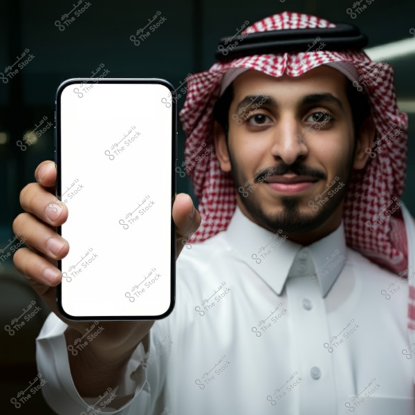 Image of a man wearing traditional Arabic clothing, including a white thobe and a red and white checkered keffiyeh, holding a smartphone with a blank white screen in his left hand while smiling. The man appears to be indoors with a dark background behind him.
